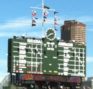 Photograph of scoreboard at Wrigley Field. In descending order:Three flagpoles showing various national league teams. The analogue clock, main board showing scores of other games, top of centerfield bleachers.